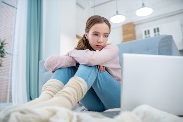 Adolescente triste en chaussettes tricotées épaisses assis sur le sol en regardant l'écran du portable.