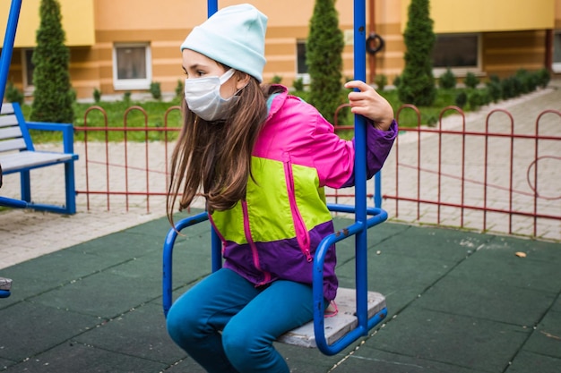 Une adolescente triste porte un masque de protection assis sur une balançoire La fille solitaire dans une aire de jeux de la ville à l'automne