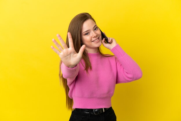 Adolescente utilisant un téléphone portable sur fond jaune isolé comptant cinq avec les doigts