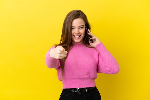 Adolescente utilisant un téléphone portable sur fond jaune isolé surpris et pointant vers l'avant