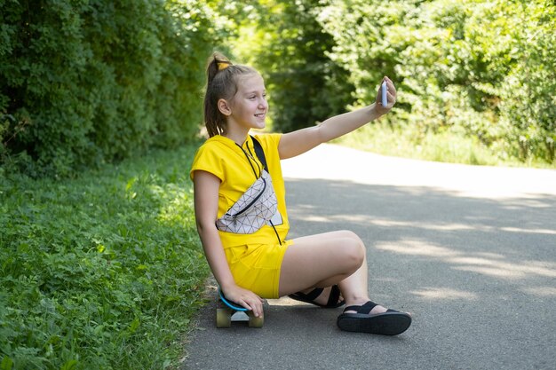 adolescente en vêtements jaunes assis sur une planche à roulettes dans un parc à l'aide d'un téléphone portable prenant un selfie