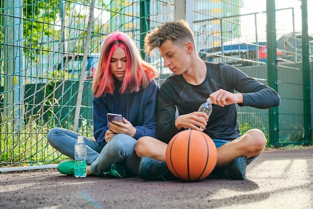 Adolescents de style de vie, garçon et fille sur un terrain de basket assis avec un ballon, parlant après le match, avec un smartphone et une bouteille d'eau. Mode de vie sain et actif, passe-temps et loisirs, concept d'adolescents