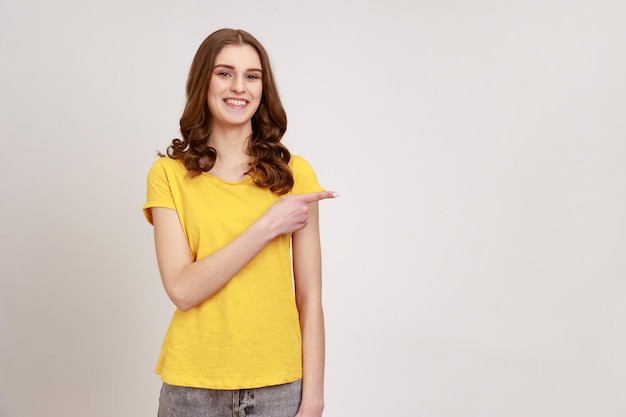 Adorable adolescente aux cheveux bouclés rouges portant un T-shirt jaune pointant vers un espace de copie vierge et souriant sincèrement, mur vide latéral pour la promotion. Tourné en studio intérieur isolé sur fond gris.