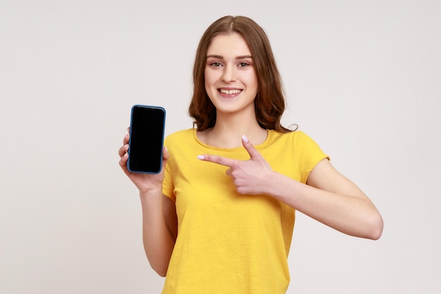 Adorable adolescente heureuse pointant son smartphone avec le doigt et souriant à la caméra publicitaire d'un appareil mobile Prise de vue en studio intérieur isolée sur fond gris