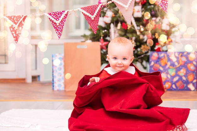 Un adorable bambin habillé en Père Noël jette un coup d'œil hors d'un sac de Noël du Père Noël dans un salon décoré avec un sapin de Noël.