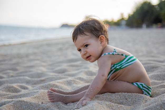 Adorable Bambin En Maillot De Bain Est Assis Sur Une Plage De Sable Au Soleil.