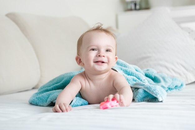 Adorable bébé allongé sous une serviette bleue sur le lit et tenant un jouet en plastique