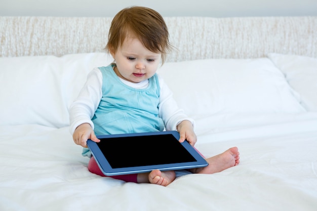 Photo adorable bébé assis sur un lit tenant une tablette informatique