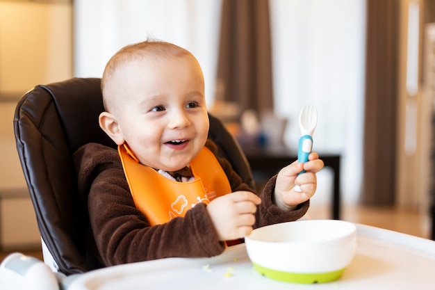 Adorable bébé mignon prenant une fourchette en plastique dans sa main et jouant avec de la nourriture assis dans la chaise haute.