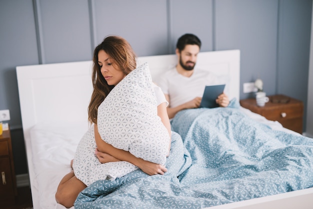 Adorable brune étreignant gros oreiller blanc tandis que jeune homme barbu vérifiant sa tablette