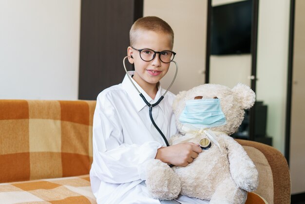 Un adorable enfant déguisé en médecin joue avec un ours en peluche tout en vérifiant sa respiration avec un st...