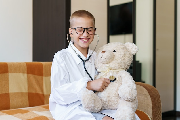 Adorable enfant habillé en docteur joue avec une peluche à la maison sur le canapé garçon heureux avec des lunettes...