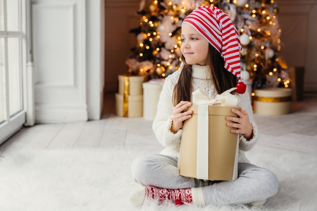 Adorable enfant de sexe féminin porte un chapeau de père Noël, des vêtements chauds, une boîte-cadeau enveloppée, s'assoit contre un arbre du Nouvel An décoré