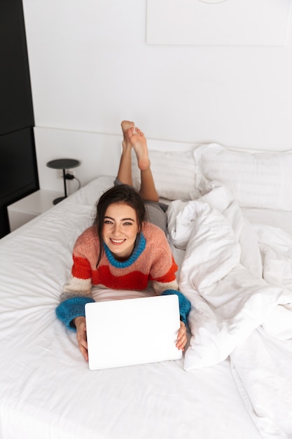 adorable femme allongée sur le lit dans l'appartement et à l'aide d'un ordinateur portable