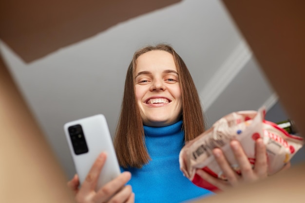 Adorable femme à l'apparence agréable regardant à l'intérieur de la boîte recevoir une commande d'épicerie tenant un colis et un téléphone portable dans les mains emballant l'aide humanitaire aux retraités et aux personnes handicapées