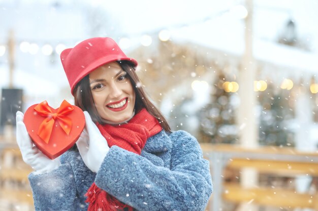Adorable femme brune en manteau d'hiver tenant une boîte-cadeau à la foire de Noël pendant les chutes de neige Espace réservé au texte