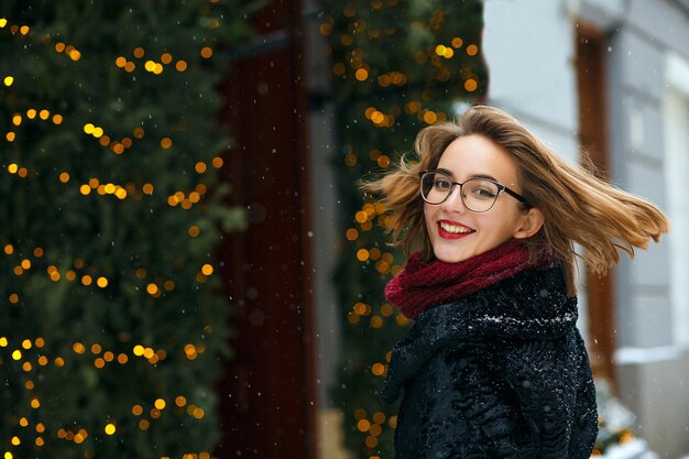 Adorable femme brune portant une écharpe et des lunettes profitant des chutes de neige. Espace libre