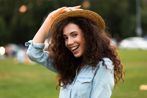 Adorable femme portant un chapeau de paille souriant, tout en marchant dans le parc verdoyant