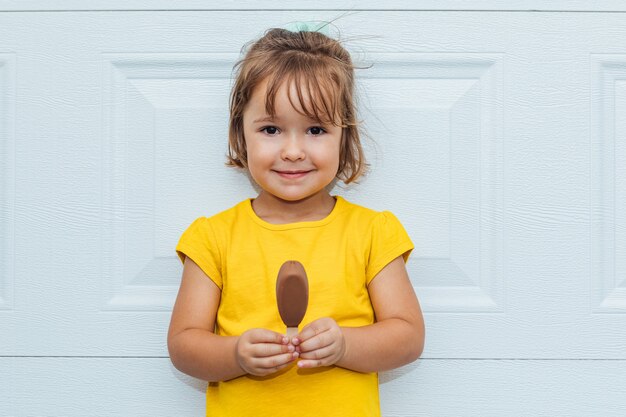 Adorable fille aux cheveux blonds, manger de la crème glacée, vêtue d'une chemise jaune appuyée sur fond blanc