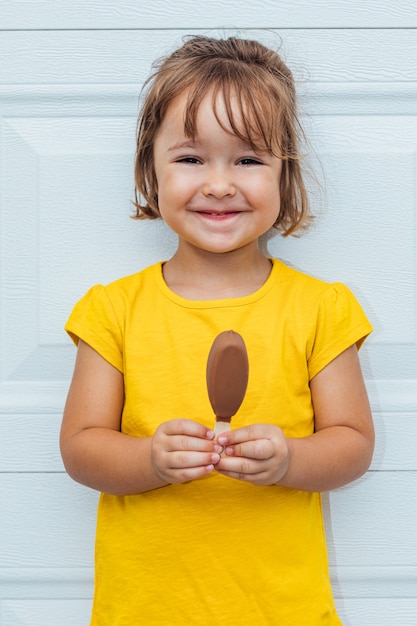 Adorable fille aux cheveux blonds, manger de la crème glacée, vêtue d'une chemise jaune appuyée sur fond blanc