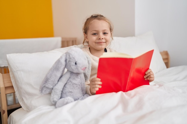 Adorable fille blonde lisant un livre assis sur le lit dans la chambre