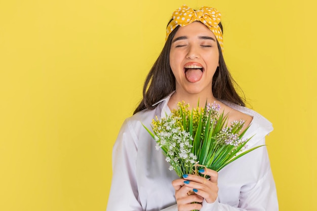 Adorable fille ferme les yeux et sort sa langue sur fond jaune avec un bouquet de fleurs Photo de haute qualité