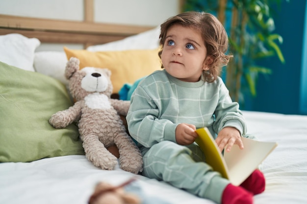 Adorable fille hispanique lisant un livre assis sur le lit dans la chambre