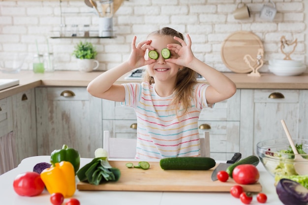 Adorable fille jouant avec du concombre frais coupé pendant la cuisson dans la cuisine à la maison