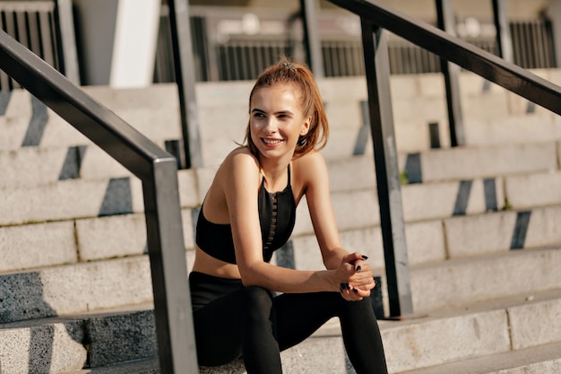 Une adorable fille souriante en uniforme de sport est assise dans les escaliers de la rue et se repose pendant l'exercice sportif Humeur joyeuse émotions vraies mode de vie sain souriant avec un beau sourire