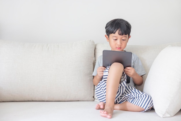 Adorable garçon asiatique heureux vêtu d'une chemise grise et d'un short rayé bleu blanc s'amuse à jouer avec sa tablette sur un canapé crème en regardant l'écran du mobile