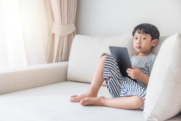 Adorable garçon asiatique heureux vêtu d'une chemise grise et d'un short rayé bleu blanc s'amuse à jouer avec sa tablette sur un canapé crème en regardant l'écran du mobile
