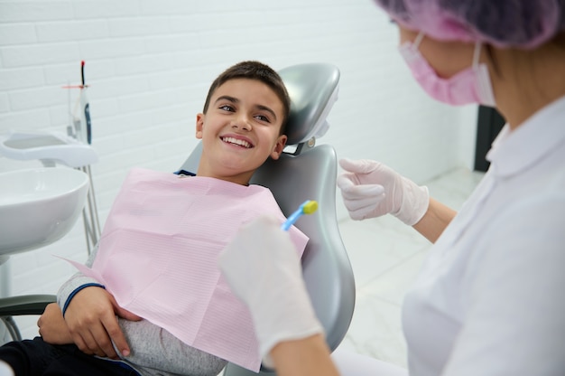 Adorable garçon assis dans une chaise de dentiste sourit avec un beau sourire à pleines dents en regardant un dentiste pédiatrique lui enseignant l'hygiène bucco-dentaire lors d'un rendez-vous dentaire dans un cabinet médical moderne et blanc