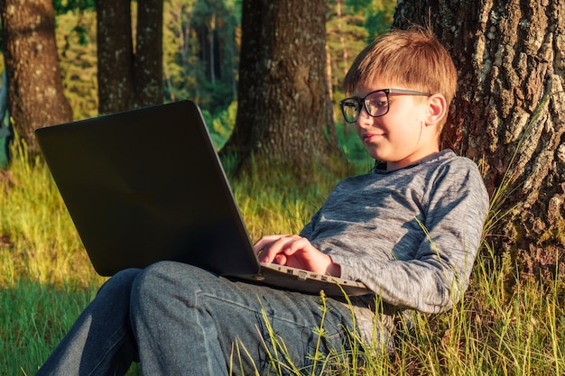 Adorable garçon junior avec des lunettes assis sur l'herbe appuyé sur un arbre dans le parc avec un ordinateur portable sur les genoux et faisant ses devoirs Élève en manches longues gris foncé et jeans tapant une tâche dans un ordinateur étudiant dans la nature