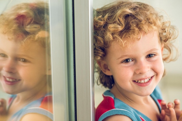 Adorable garçon souriant aux cheveux blonds bouclés regardant loin tout en étant assis près de la fenêtre avec réflexion sur le balcon d'un appartement résidentiel