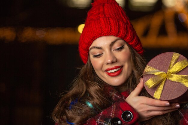 Adorable jeune femme au chapeau rouge recevant un cadeau dans une boîte à la foire de Noël le soir. Espace libre