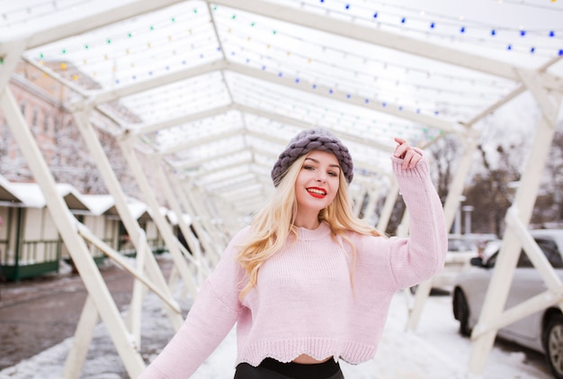 Adorable jeune femme au maquillage lumineux portant un bonnet et un pull en tricot, posant dans la rue en hiver. Espace pour le texte
