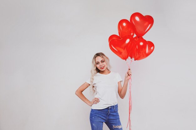 Adorable jeune fille blonde en jeans et t-shirt blanc posant avec des ballons en forme de coeur sur fond blanc concept de la Saint-Valentin