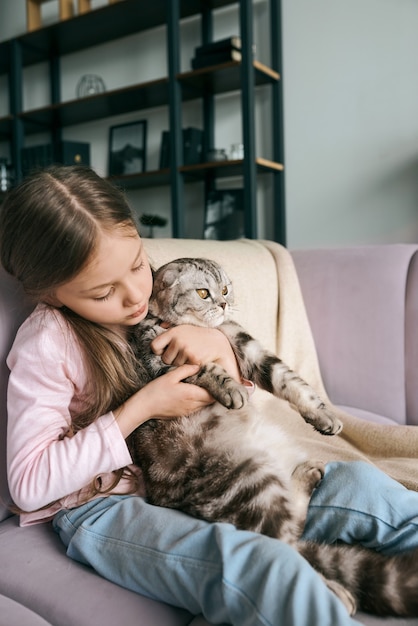 Adorable jolie fille souriante étreignant un chat gris écossais sur le canapé à la maison