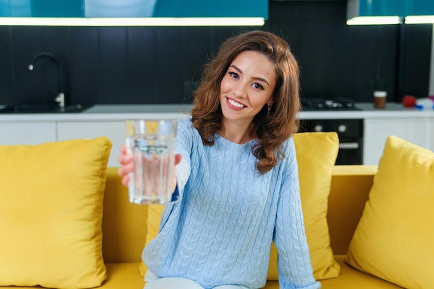 Adorable joyeuse femme aux cheveux ondulés regardant la caméra avec un verre d'eau fraîche sur la cuisine