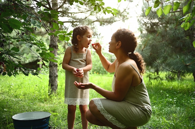 Adorable mère nourrit sa fille avec des cerises cueillies dans le jardin. Maman et sa fille s'habillent de la même façon lorsqu'elles cueillent des cerises en été. Maternité, enfance, relations familiales.