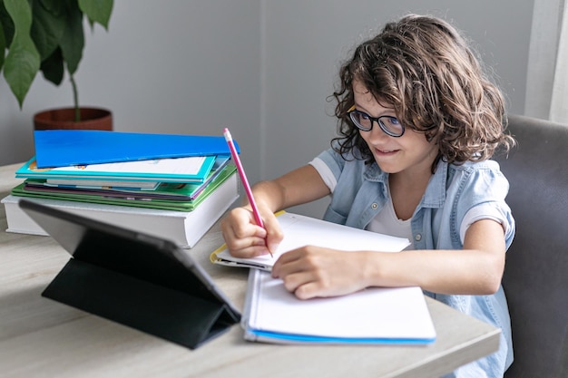 Adorable petit écolier avec des lunettes étudiant en ligne depuis un enfant à la maison à l'aide d'une tablette numérique