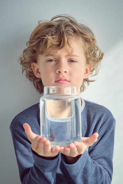 Adorable petit enfant aux cheveux blonds bouclés et aux yeux marron portant un pull bleu montrant une bouteille en verre transparent d'eau propre et regardant la caméra sur fond blanc