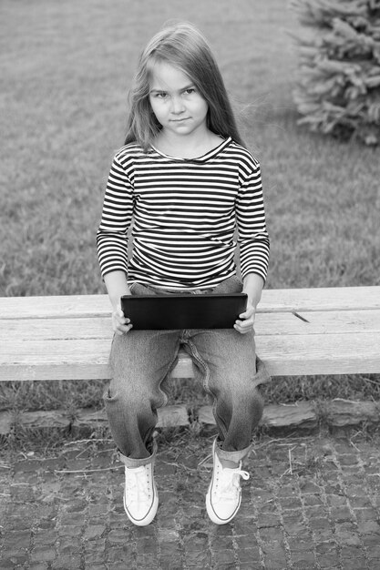 Adorable petit enfant dans un style décontracté avec un look intelligent utilise une tablette moderne assis sur un banc de parc à l'extérieur de l'ordinateur