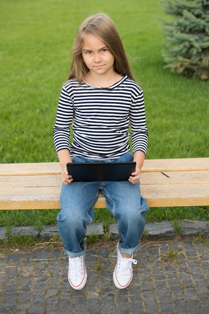 Adorable petit enfant dans un style décontracté avec un look intelligent utilise une tablette moderne assise sur un banc de parc à l'extérieur, un ordinateur.