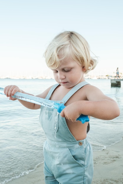 Adorable petit enfant explorant un récipient avec des bulles de savon au bord de la mer avec une côte de lavage à l'eau par temps ensoleillé