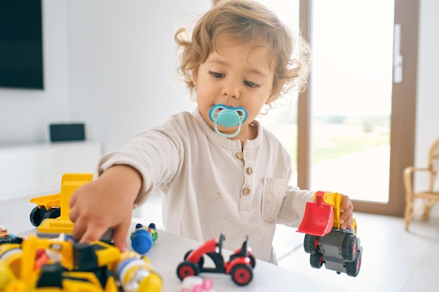 Adorable petit enfant avec une sucette jouant avec des jouets en plastique colorés à la maison dans une journée ensoleillée sur un fond flou