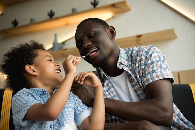 Adorable petit garçon afro-américain tenant une cuillère et nourrissant un père heureux incafe