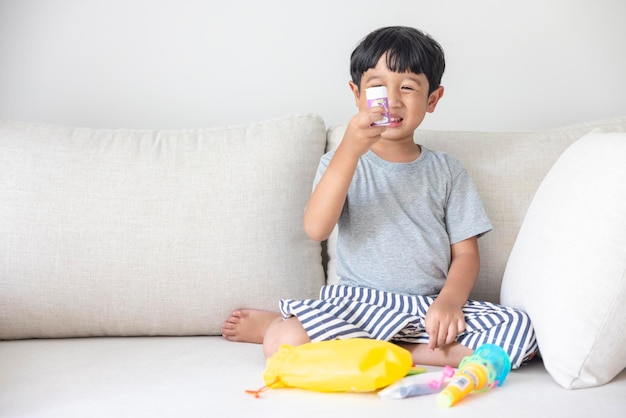 Adorable petit garçon asiatique heureux vêtu d'une chemise grise et d'un short rayé bleu blanc assis sur un canapé crème sourit joyeusement choisit ses jouets intentionnellement