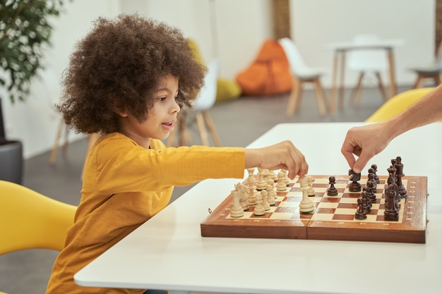 Adorable Petit Garçon Faisant Un Geste En Jouant Aux échecs Avec Un Adulte Assis à La Table à L'intérieur