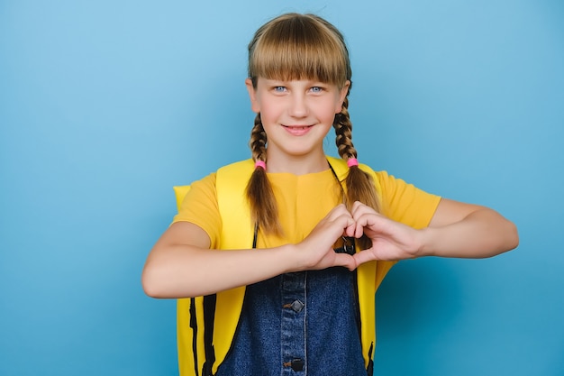 Adorable petite écolière mignonne montrant le geste du cœur avec les mains, souriante en regardant la caméra, porte un sac à dos jaune, posant isolée sur fond de couleur bleue en studio. Retour au concept de l'école